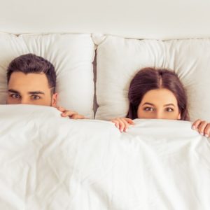 youmg couple in bed peeping over the bed sheet
